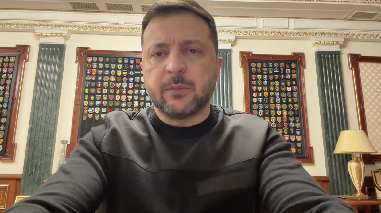 Чоловік у темному одязі на фоні стін із військовими шевронами у рамках та зеленими колонами.