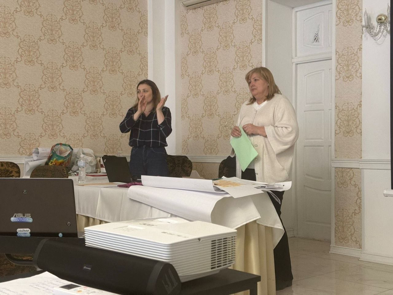 Дві жінки проводять презентацію або тренінг у світлій залі. Одна жінка виступає перед аудиторією, тримаючи в руках зелений аркуш паперу, інша стоїть поруч і жестикулює. На столі перед ними лежать папери, ноутбук та стоїть проектор.