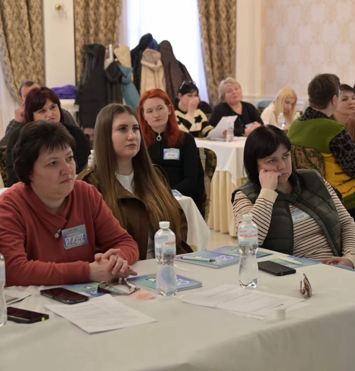 Група жінок сидить за столами під час семінару або тренінгу. Учасниці уважно слухають спікера, на столах перед ними лежать робочі матеріали, блокноти та пляшки з водою. На задньому плані видно інших слухачів та інтер’єр зали з бежевими стінами та шторами.