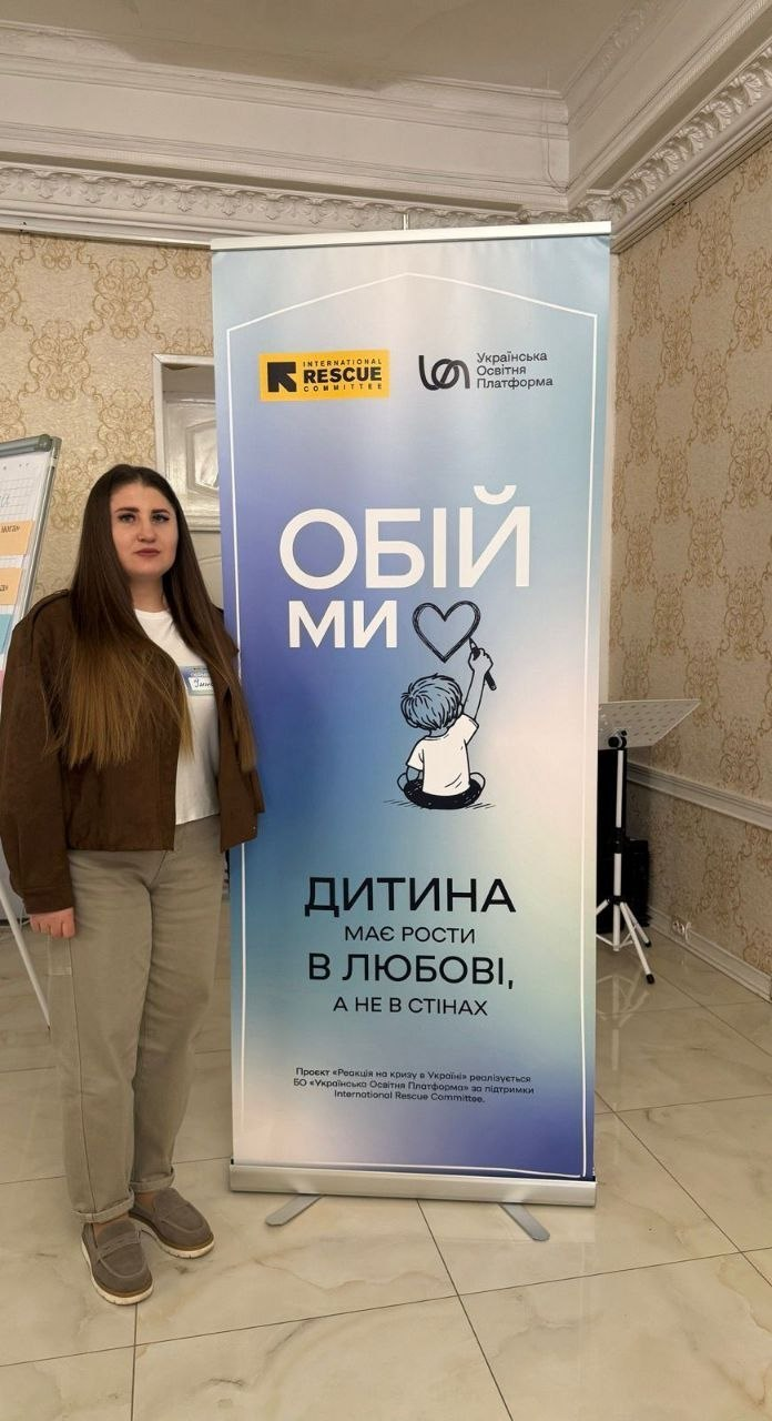Жінка стоїть поруч із інформаційним стендом. На банері зображено малюнок дитини, що малює серце, та напис: «Обійми. Дитина має рости в любові, а не в стінах». У верхній частині стенда розміщено логотипи «International Rescue Committee» та «Українська освітня платформа».