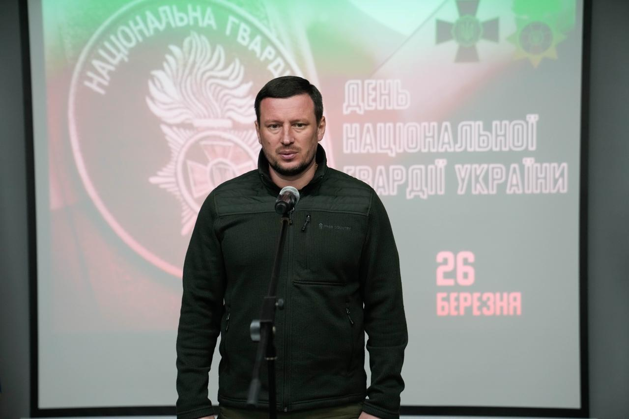 Чоловік у тактичному одязі виступає біля мікрофона. На фоні розташований екран із символікою та написом про День Національної гвардії України (26 березня).