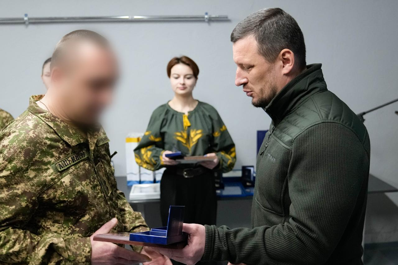 Чоловік у тактичному одязі вручає нагороду військовослужбовцю. На задньому плані стоїть жінка у вишиванці, яка тримає наступну відзнаку. Обличчя військового розмито.