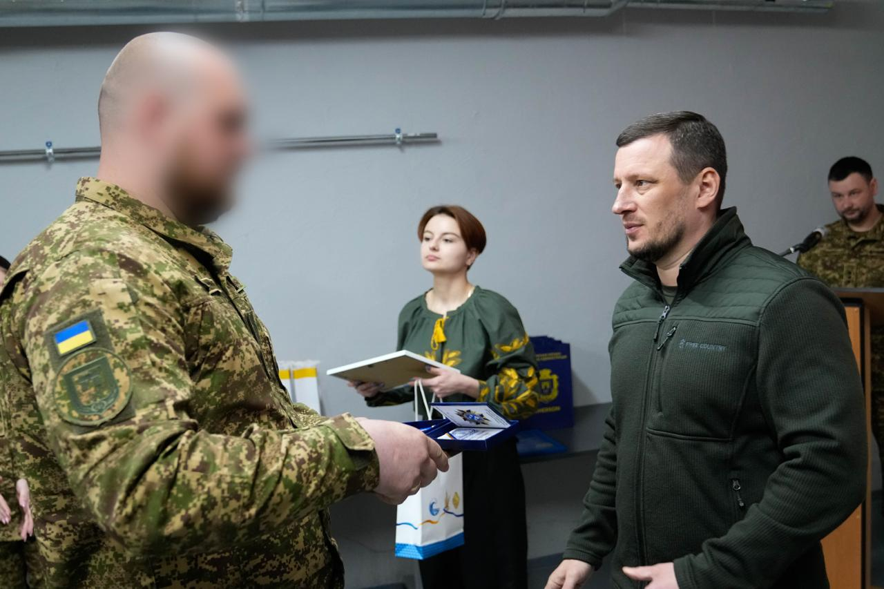 Чоловік у тактичному одязі спілкується з військовослужбовцем під час нагородження. На задньому плані стоїть жінка у вишиванці з нагородами в руках. Обличчя військового на передньому плані розмито.