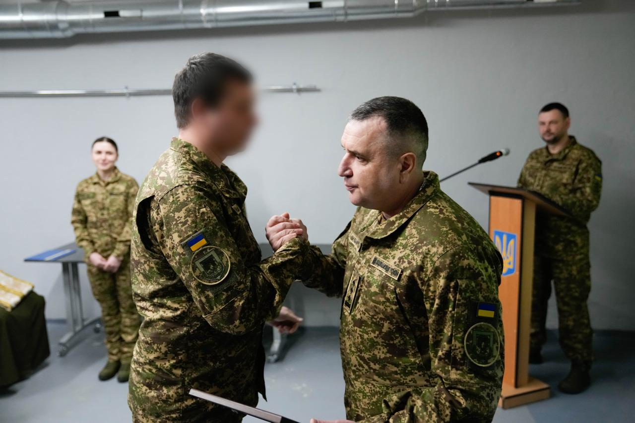 Військовий керівник тисне руку військовослужбовцю під час нагородження. На задньому плані стоїть жінка у формі та чоловік за трибуною з емблемою Національної гвардії України. Обличчя одного з військових розмито.