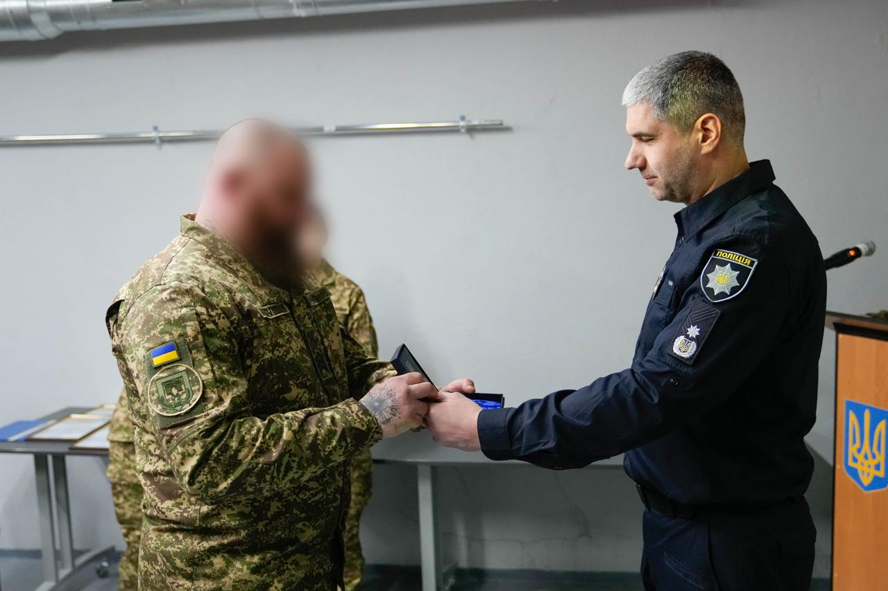 Представник поліції вручає нагороду військовослужбовцю. Поліцейський у темно-синій формі передає відкритий футляр із відзнакою військовому у камуфляжі. Обличчя військовослужбовця розмито.