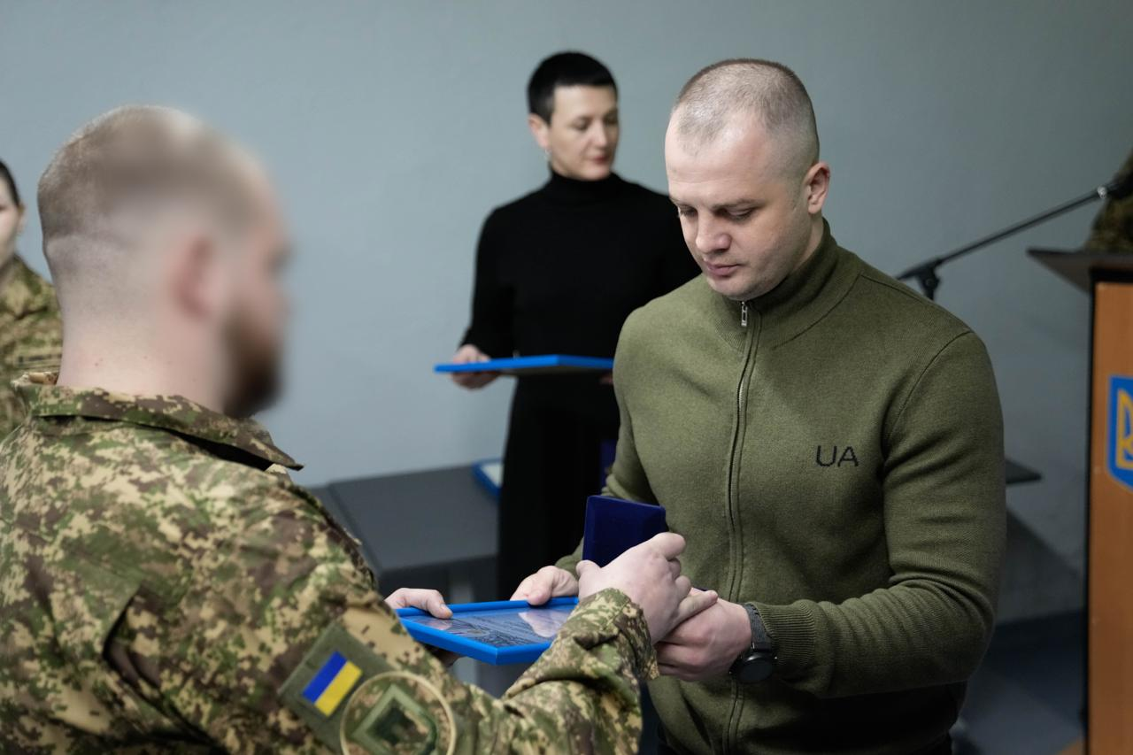 Військовослужбовець у камуфляжі вручає нагороду та грамоту чоловікові в оливковій кофті. На задньому плані стоїть жінка в чорному одязі, яка тримає ще одну грамоту. Обличчя військового на передньому плані розмито.