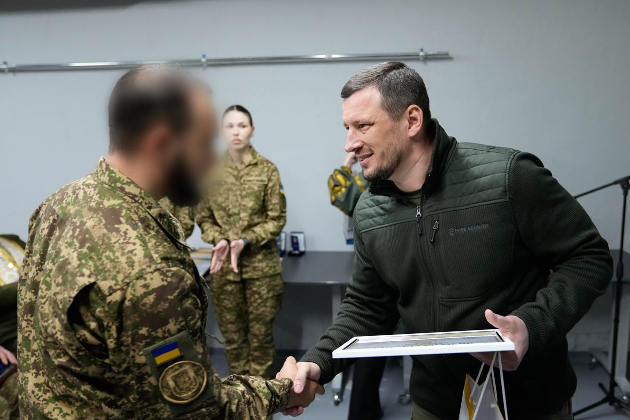 Чоловік у темно-зеленій кофті тисне руку та вручає грамоту військовослужбовцю. На задньому плані стоїть жінка у військовій формі. Обличчя військового на передньому плані розмито.