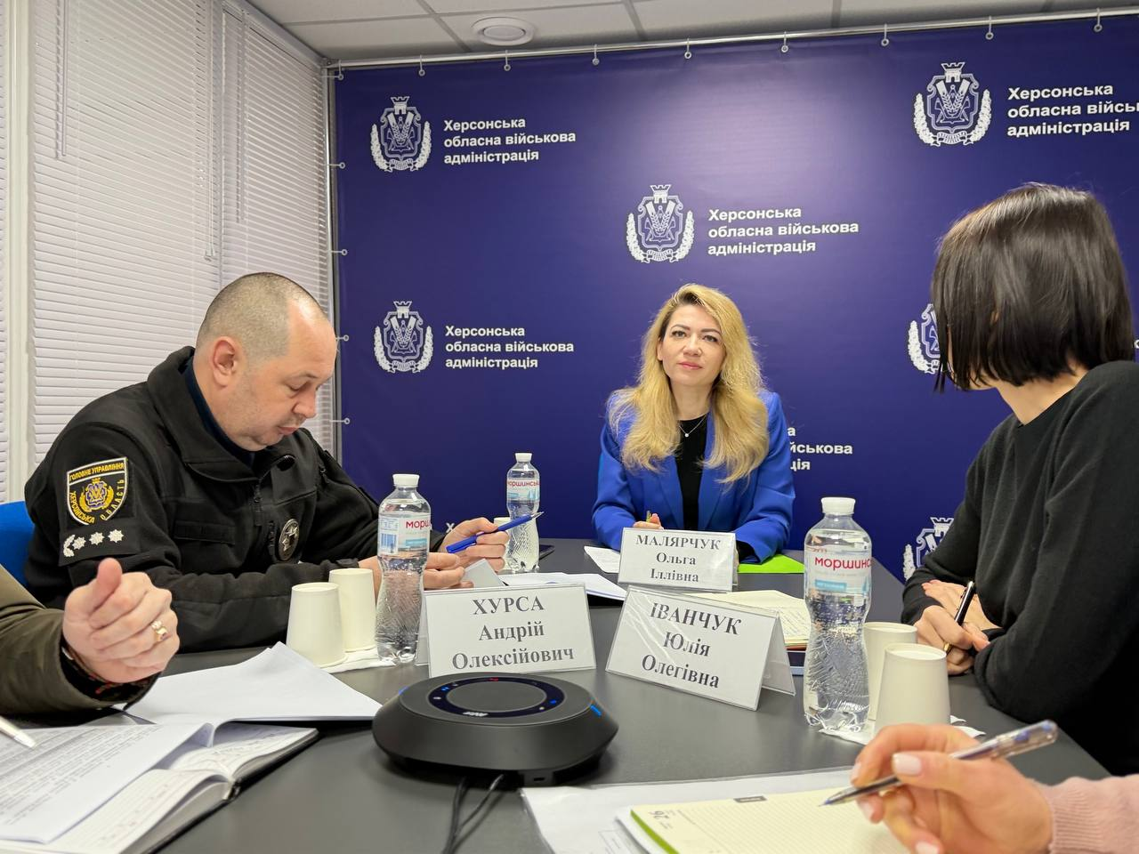 Робоча зустріч посадовців у офіційному кабінеті. Кілька осіб сидять за столом, на якому лежать документи, таблички з іменами, пляшки води та пристрій для зв'язку. На задньому плані — брендований синій банер із гербом та офіційною назвою установи.