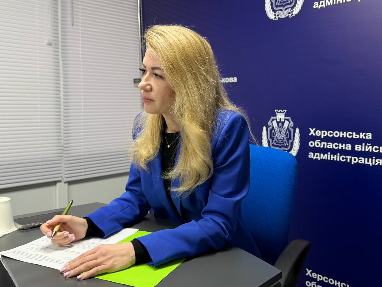 Жінка в яскраво-синьому піджаку сидить за робочим столом. Вона тримає ручку над документами на зеленій папці та дивиться вбік. На задньому плані — темно-синій банер із гербом та офіційним написом установи.