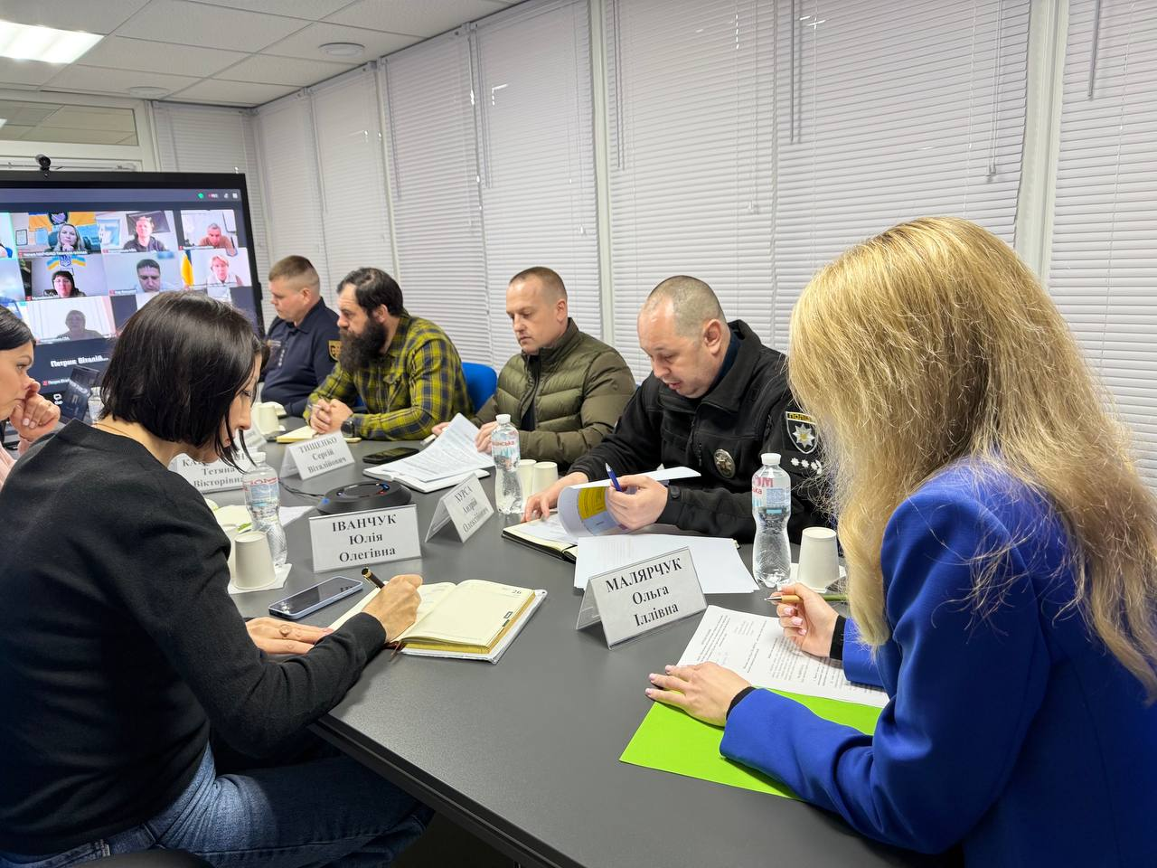 Робоча нарада в офіційному приміщенні з використанням відеозв'язку. Учасники в цивільному та форменому одязі сидять за столом із документами та табличками. На задньому плані розташований великий екран, на якому відображено вікна учасників онлайн-конференції.