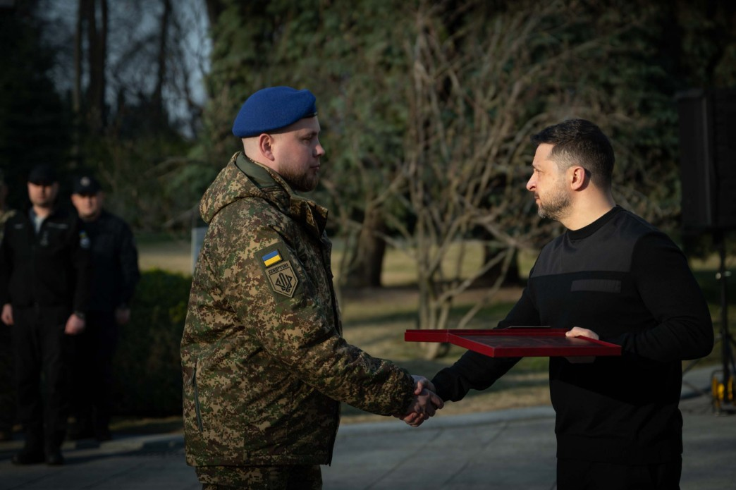Чоловік у чорному одязі тисне руку військовослужбовцю у камуфляжі та синьому береті. У руках він тримає червоний футляр із нагородою. На задньому плані видно дерева та інших людей у формі.