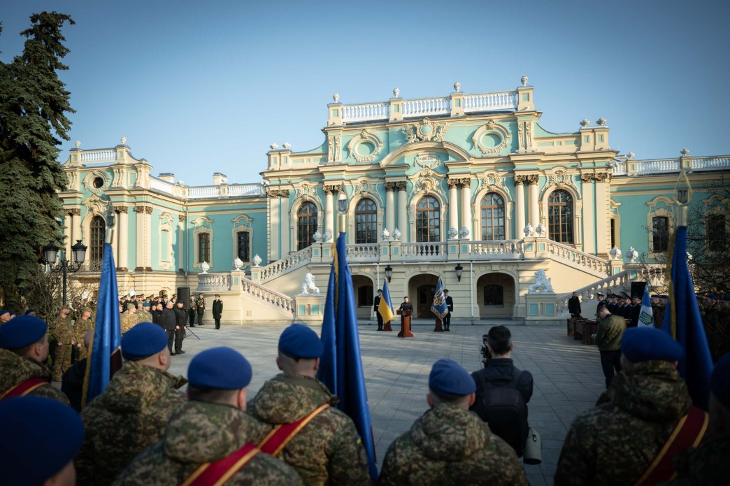 Урочиста церемонія за участю військовослужбовців на площі перед Маріїнським палацом. На передньому плані — група військових у синіх беретах, що стоять спиною до камери. Вдалині, перед центральним входом до палацу, за трибуною виступає особа поруч із державним прапором України та стягом Національної гвардії.
