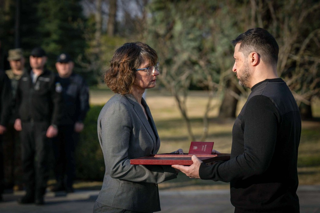 Чоловік у чорному одязі вручає нагороду жінці в сірому піджаку на офіційній церемонії просто неба. На задньому плані стоять люди у форменому одязі, а навколо видно дерева.