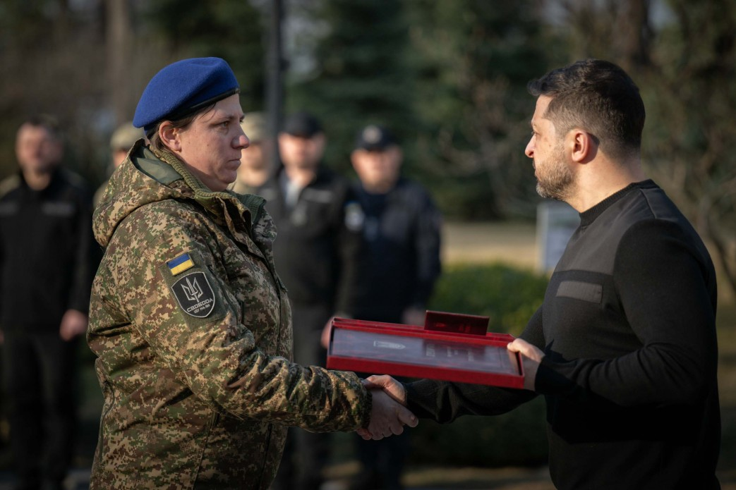 Чоловік у чорному одязі тисне руку та вручає нагороду жінці-військовослужбовиці. Вона одягнена у камуфляжну куртку та синій берет, на плечі видно шеврон із гербом України. Нагородження відбувається на вулиці на фоні інших людей у формі та дерев.