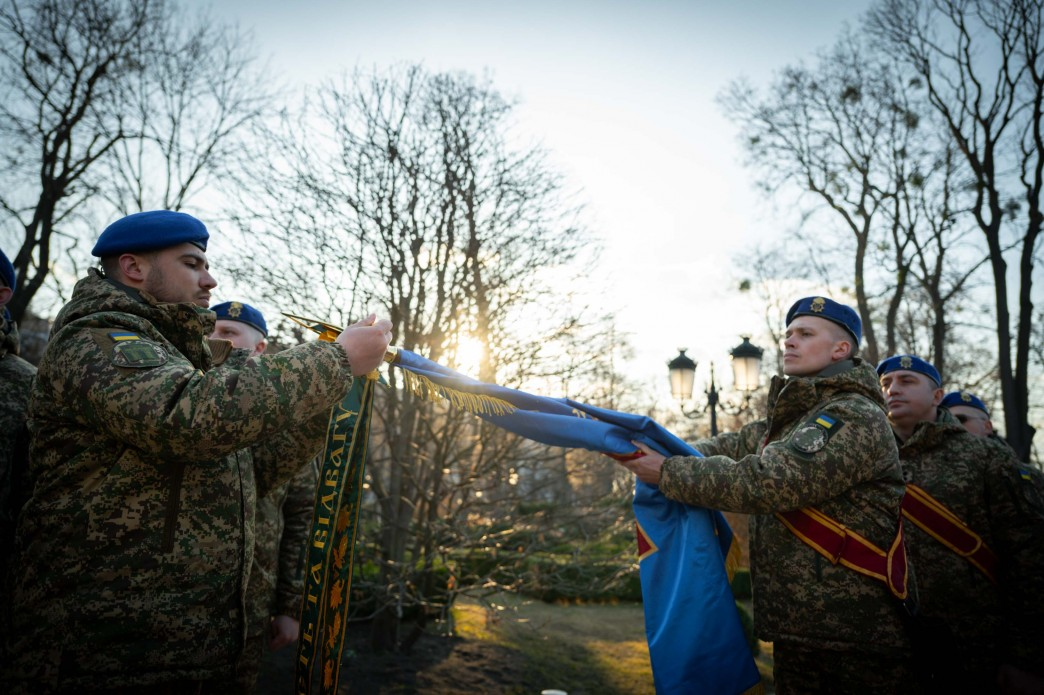 Військовослужбовці Національної гвардії України під час урочистого ритуалу з прапором. Група гвардійців у камуфляжній формі та синіх беретах тримає розгорнуте бойове знамено зі стрічками. Дія відбувається на вулиці на фоні дерев та призахідного сонця.