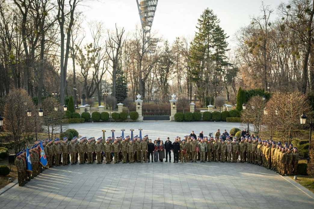 Групове фото військовослужбовців та цивільних осіб на площі перед Маріїнським палацом. Велика кількість гвардійців у камуфляжній формі та синіх беретах вишикувана півколом. У центрі стоять офіційні особи, а збоку розташована група з прапорами. На задньому плані видно парк, огорожу та верхівку металевої конструкції.