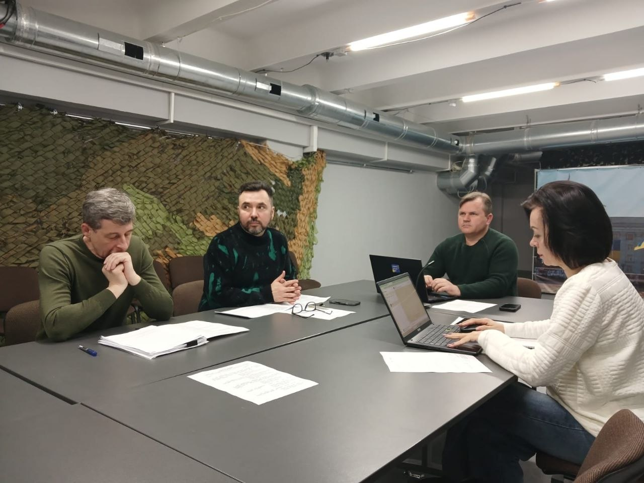 Четверо людей сидять за великим столом у робочому приміщенні. Вони зосереджено працюють із ноутбуками та паперами. На стіні позаду — маскувальна сітка.