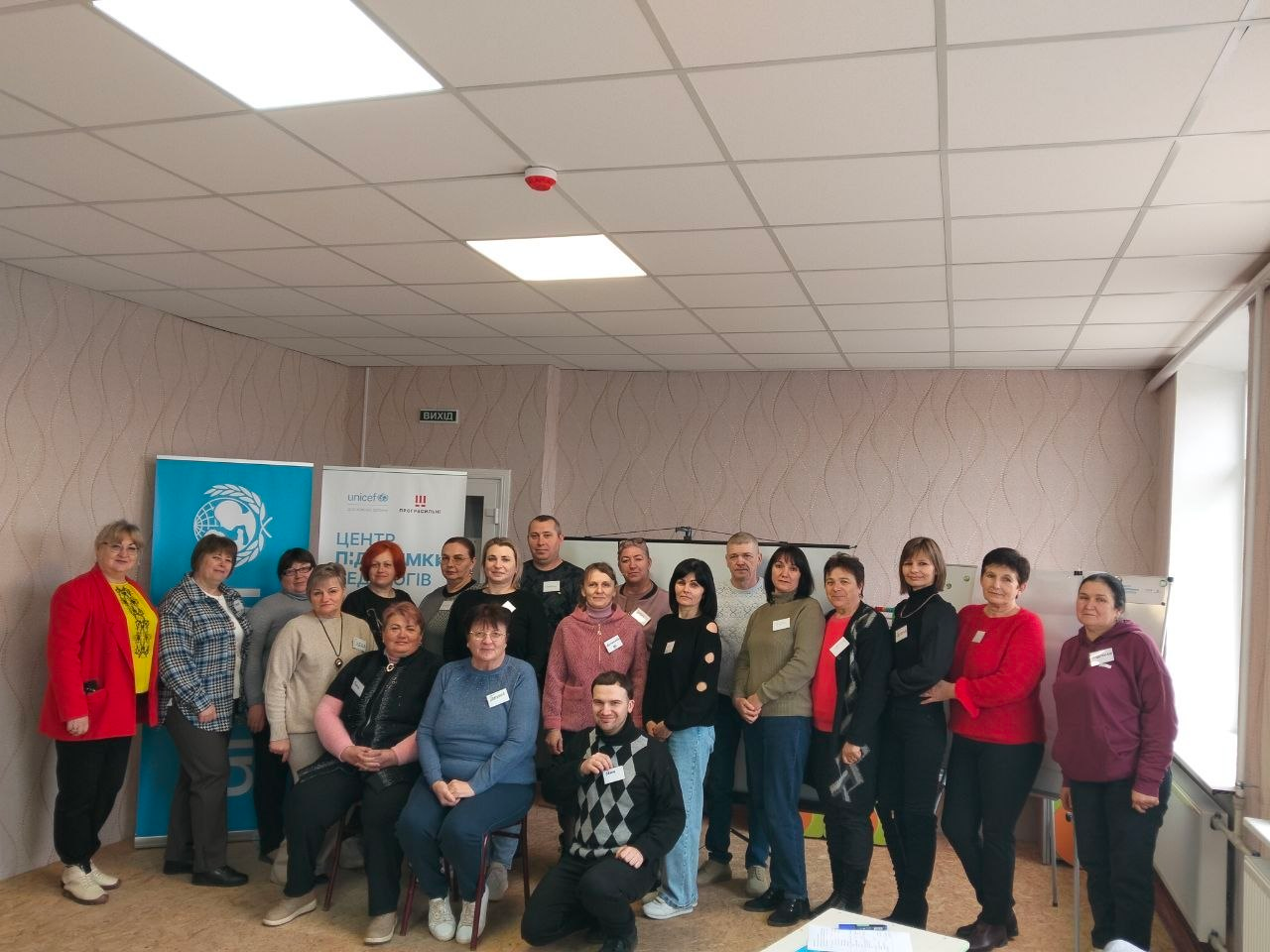 Групове фото близько двадцяти людей у світлому офісному приміщенні. Учасники стоять і сидять на фоні інформаційних банерів із логотипами та написом «Центр підтримки...». Позаду — фліпчарт та вікно.
