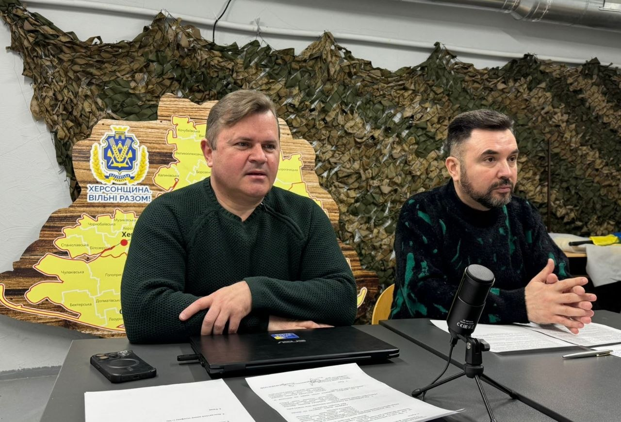 Двоє чоловіків за столом під час засідання на фоні маскувальної сітки та дерев’яної карти Херсонщини. Перед ними — мікрофон, ноутбук і документи.