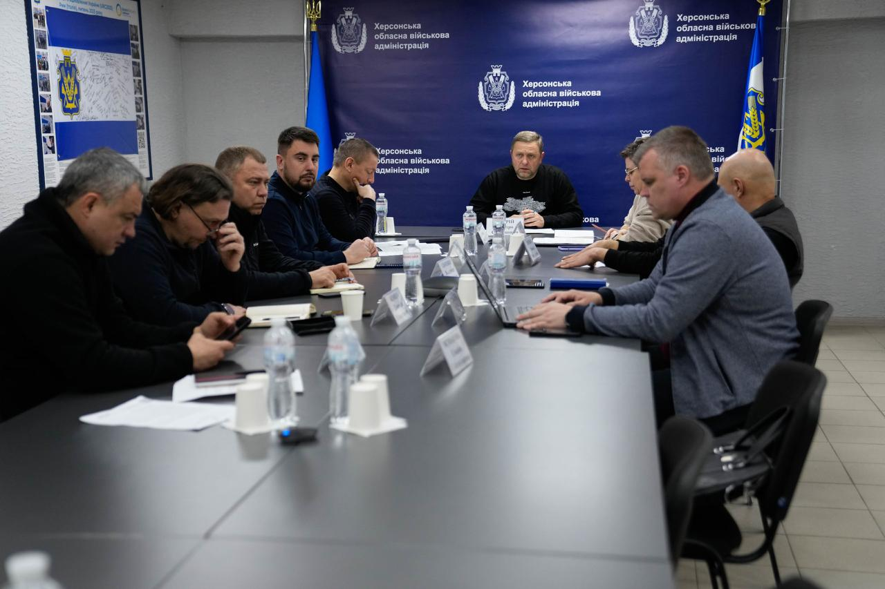 Офіційна робоча нарада у кабінеті. Група учасників сидить за довгим столом із документами, ноутбуками та водою. На задньому плані — синій банер із державною символікою та прапори України.