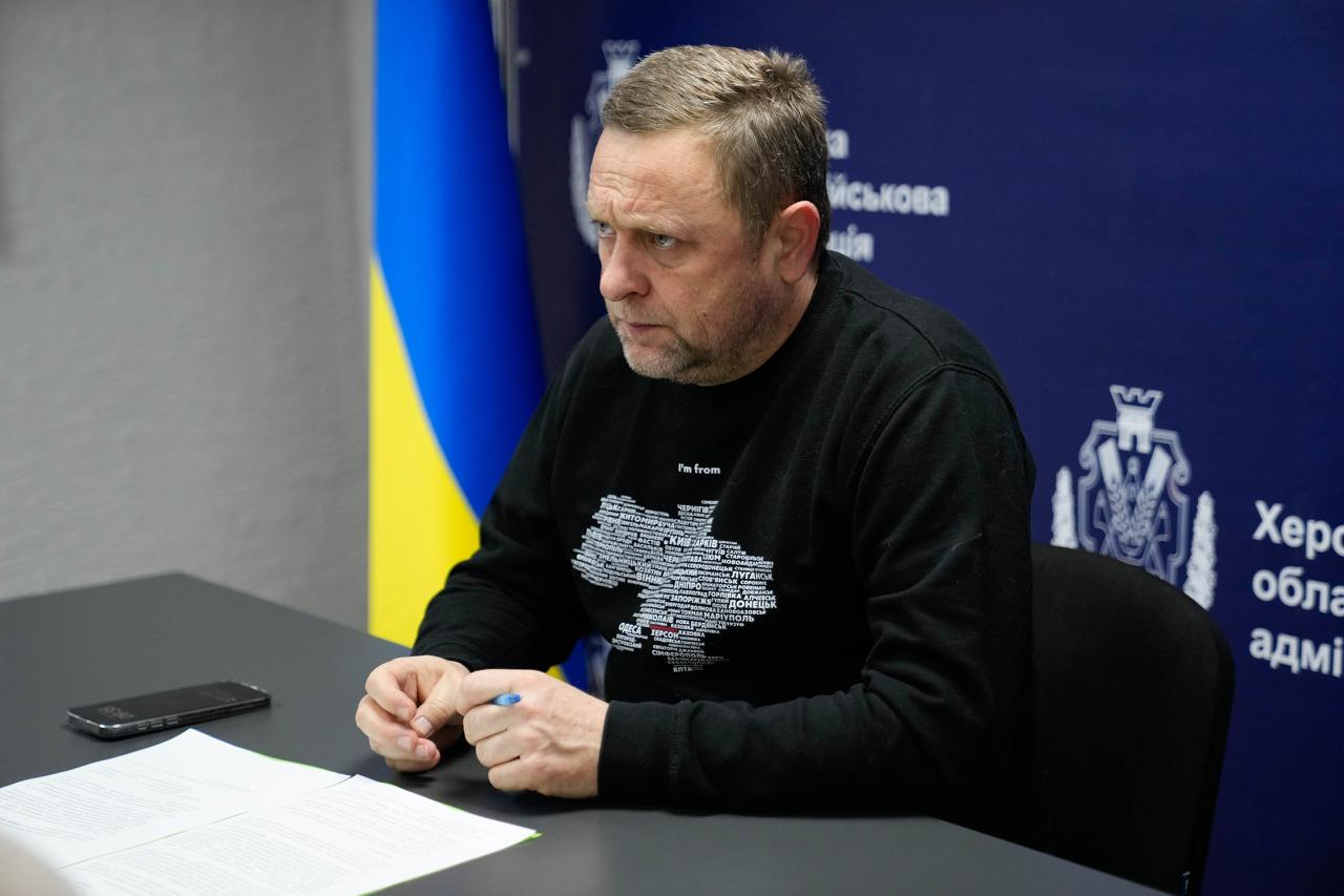 Чоловік у чорному світшоті з картою України сидить за робочим столом із документами та ручкою. На фоні — синій банер із державною символікою та Державний Прапор України.