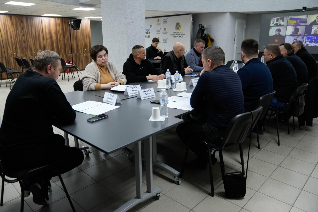 Група людей на робочій нараді за великим Т-подібним столом із табличками та документами. На задньому плані — екран із учасниками відеоконференції та банер медіацентру.