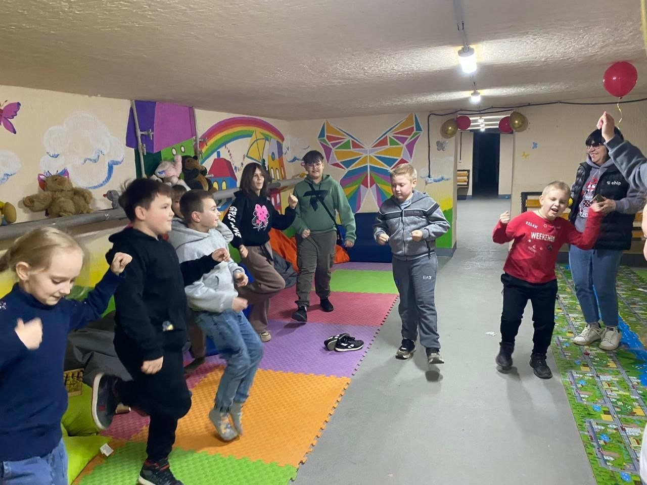 Група дітей різного віку весело танцює та грається в ігровій кімнаті. Приміщення оздоблене яскравими малюнками на стінах, м'якими іграшками та кольоровим покриттям на підлозі.