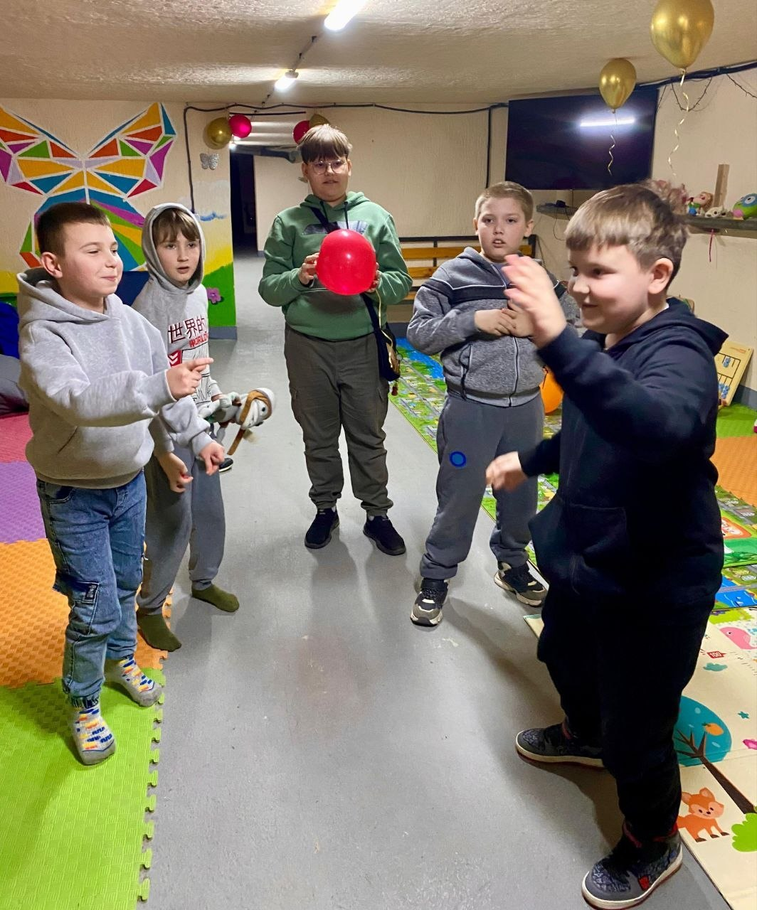 П’ятеро хлопчиків граються в ігровій кімнаті або укритті. Один із них тримає червону повітряну кульку, інші спілкуються та жестикулюють. Стіни приміщення оздоблені яскравими малюнками, на підлозі лежить кольоровий дитячий килимок-пазл.