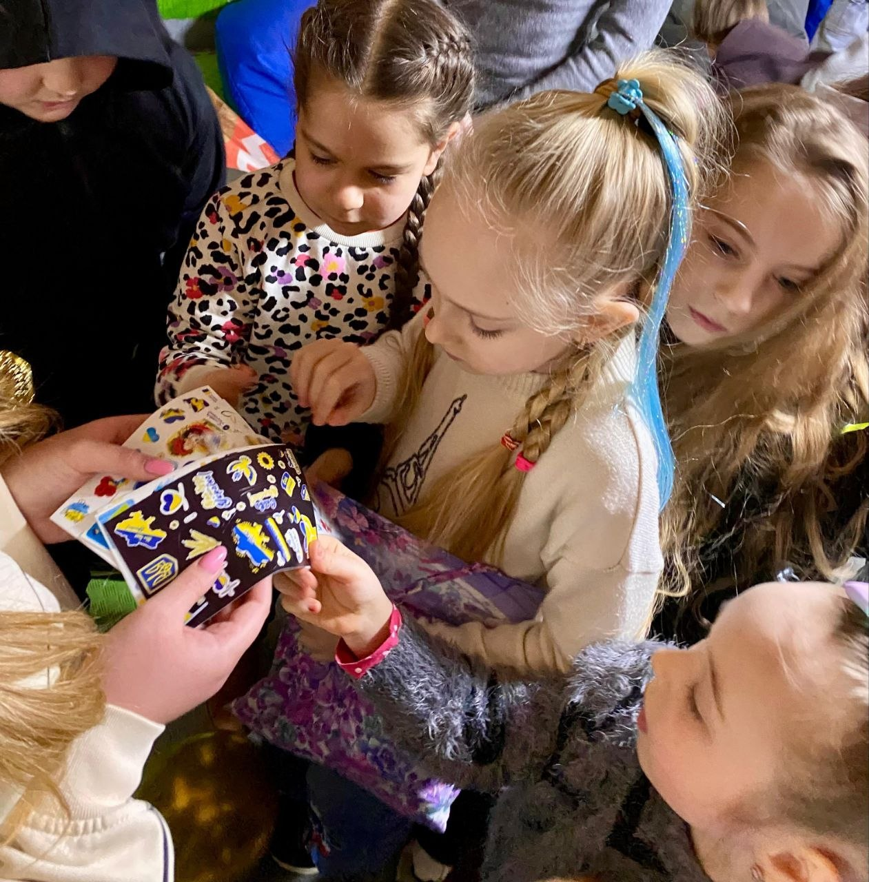 Група дітей зосереджено розглядає аркуші з патріотичними українськими наклейками. На стікерах зображено державну символіку, серця та літаки в жовто-блакитних кольорах.