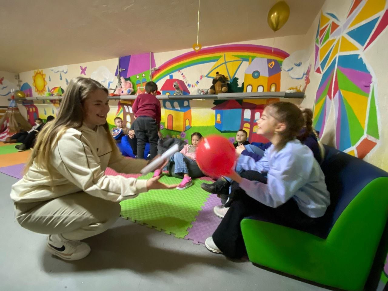 Група дітей та дівчина-аніматор весело проводять час в ігровій кімнаті з розмальованими стінами. Дівчина сидить навпочіпки, простягаючи руки до дівчинки з червоною повітряною кулькою. У кімнаті багато іграшок, м’яких крісел та кольорове покриття на підлозі.