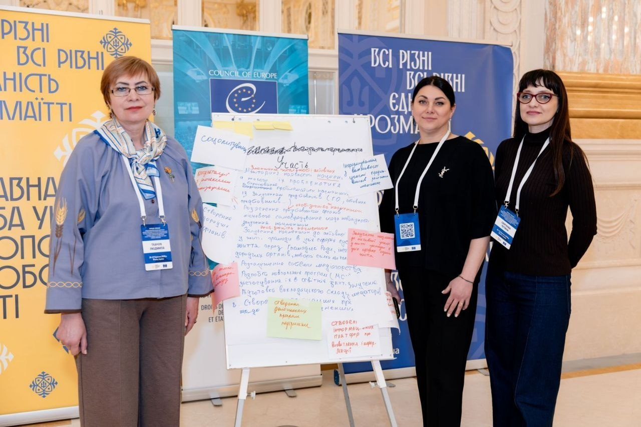 Три учасниці офіційного заходу стоять поруч із фліпчартом, на якому розміщено рукописні тези та кольорові стікери. На задньому плані — банери з символікою Ради Європи та гаслом «Всі різні, всі рівні».