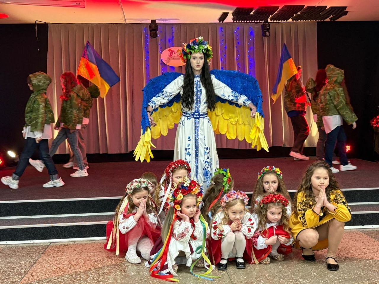 Сценічна постановка на патріотичну тематику. У центрі стоїть дівчина в білій вишиванці, квітковому вінку та з великими жовто-блакитними крилами. Перед нею на підлозі сидять маленькі діти у вінках, а на задньому плані видно учасників із прапорами України та в камуфляжному одязі.