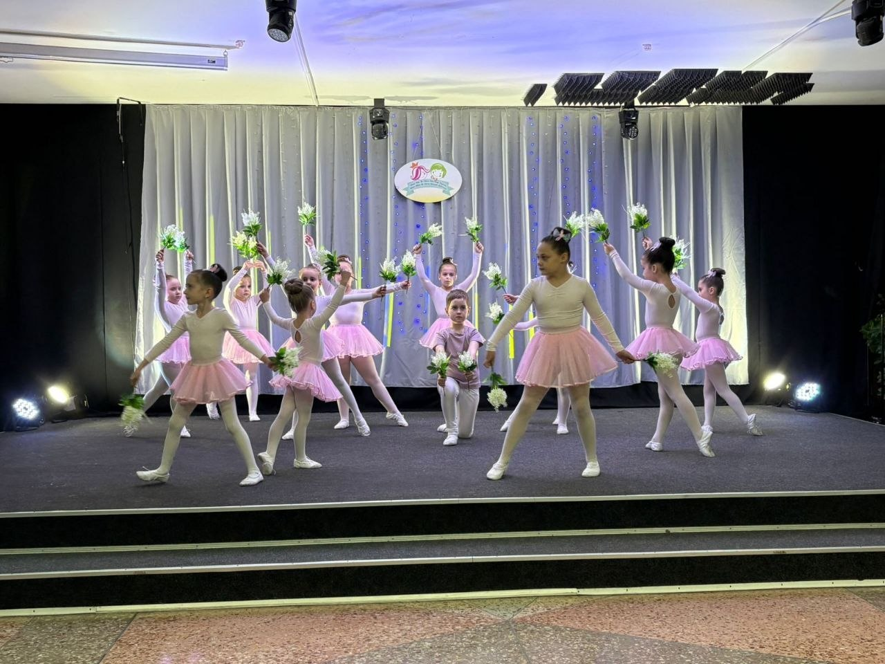 Група дітей у рожевих балетних пачках та білих костюмах виступає на сцені. Учасники танцюють, тримаючи в руках білі квіти. На задньому плані — світла завіса з емблемою заходу та сценічне освітлення.