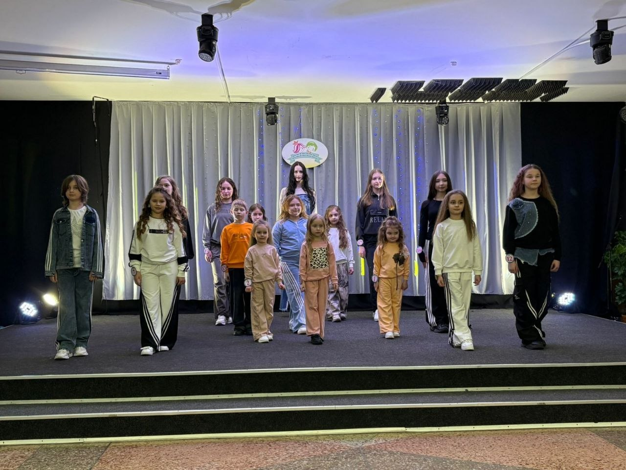 Група дітей та підлітків позує на сцені під час виступу або конкурсу. Учасники одягнені в сучасний повсякденний одяг та спортивні костюми. На задньому плані — світла штора з невеликою емблемою по центру та сценічне освітлення.