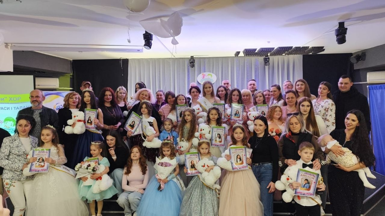 Групове фото учасників дитячого конкурсу на сцені. Діти у святкових сукнях та костюмах тримають дипломи і м’які іграшки. На фоні — банер конкурсу краси та талантів і світла завіса.