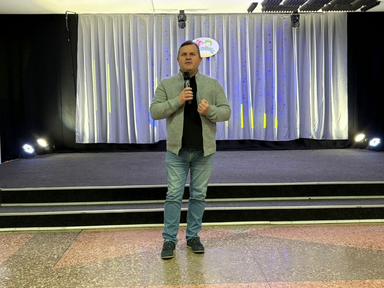 Чоловік у сірому кардигані та джинсах виступає з мікрофоном на сцені. На задньому плані — світла завіса з невеликим логотипом по центру та сценічне освітлення.