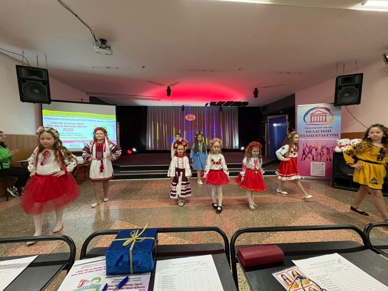 Група дівчаток у яскравих вишиванках та вінках виступає на сцені під час дитячого фестивалю. На задньому плані — екран із назвою заходу «Маленька пані та Містер Херсон — 2026» та банер Херсонського обласного палацу культури. На передньому плані — стіл журі з протоколами та подарунком.
