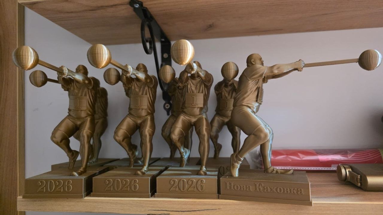 Ряд золотистих статуеток на дерев'яній полиці, що зображують атлета в момент метання молота. На підставках фігурок видно написи «2026» та «Нова Каховка».