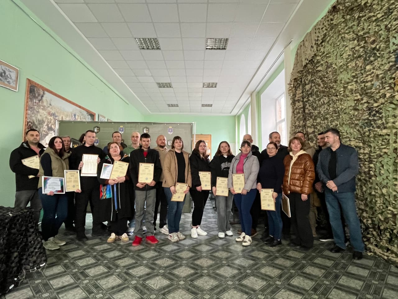 Групове фото людей у приміщенні, які тримають у руках почесні грамоти та подяки. Учасники стоять у два ряди на фоні стіни, частина якої завішена маскувальною сіткою. Атмосфера заходу урочиста та офіційна.