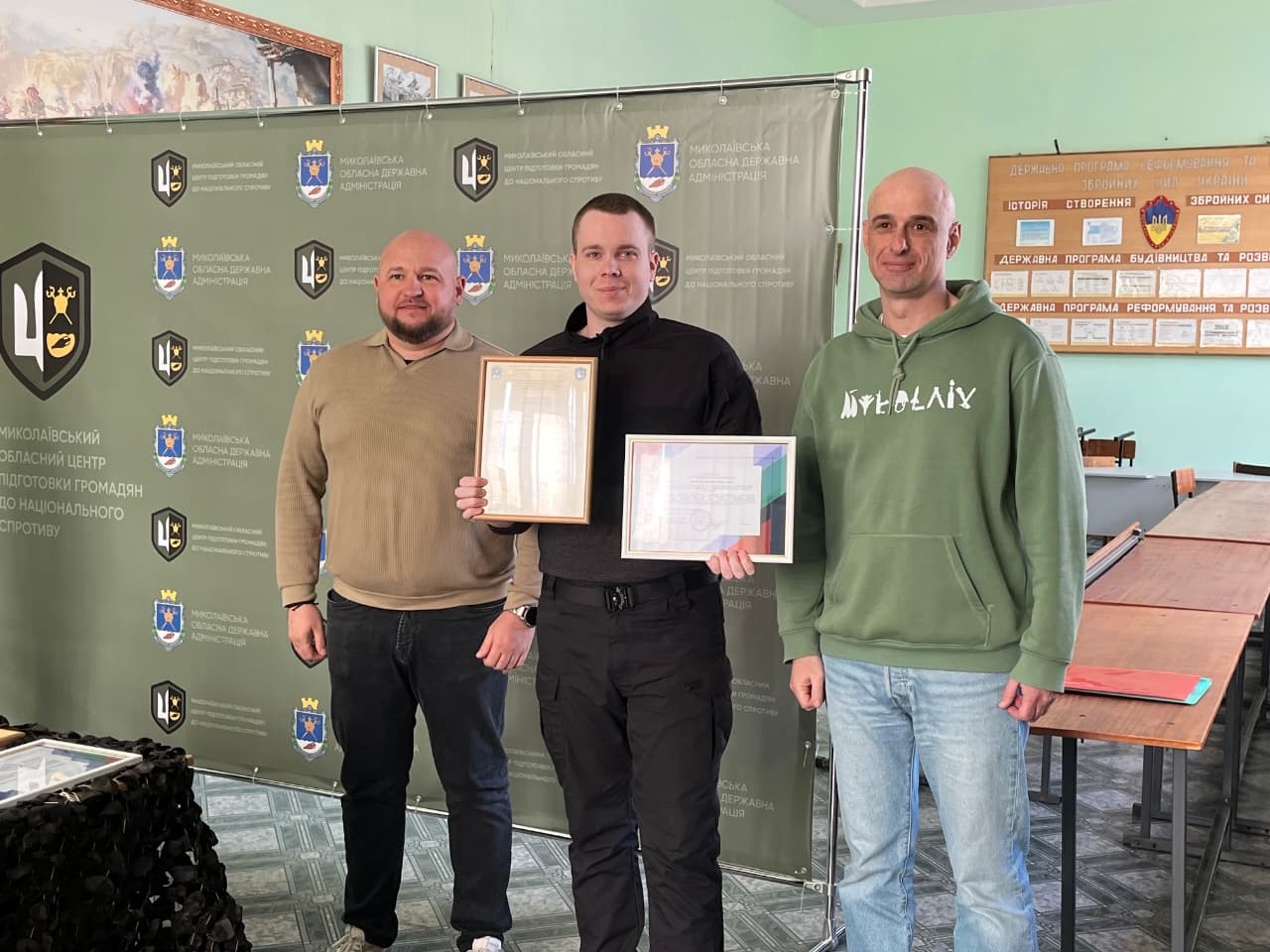 Троє чоловіків у приміщенні на фоні офіційного банера з гербами та інформаційного стенда. У центрі — чоловік середньої статури з короткою зачіскою, одягнений у чорну форму, який тримає в руках дві почесні грамоти в рамках. Атмосфера події — офіційне нагородження.