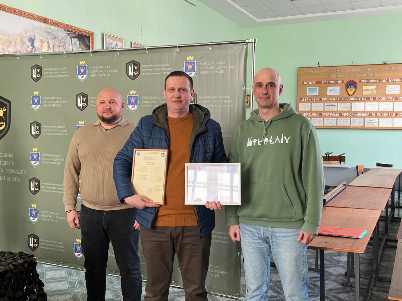 Троє чоловіків у повсякденному одязі стоять у приміщенні на фоні офіційного банера. У центрі — чоловік середньої статури з коротким русявим волоссям, одягнений у темно-синю куртку та джемпер цегляного кольору; він тримає в руках дві почесні грамоти в рамках. Атмосфера події — урочисте нагородження.