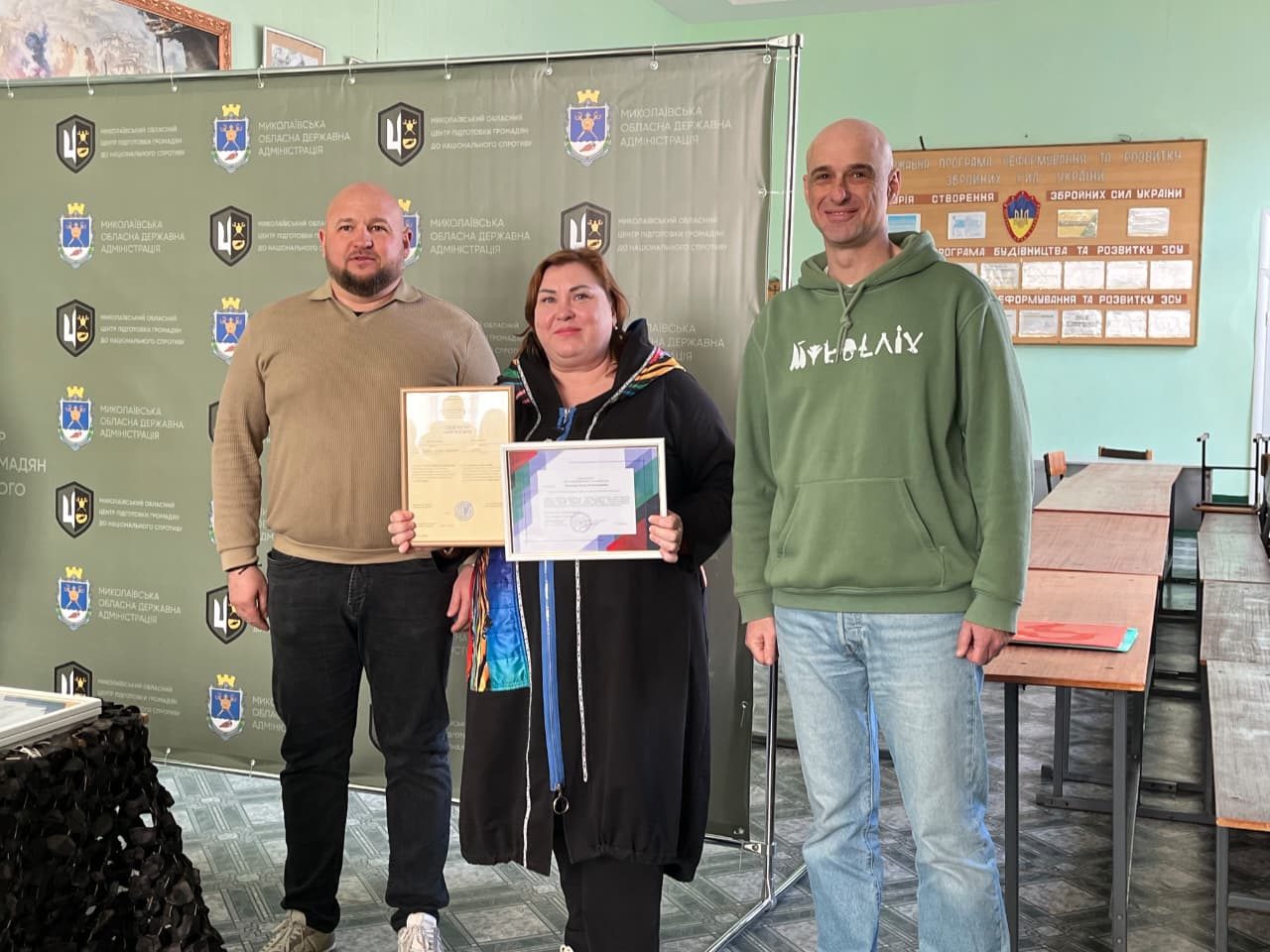 Троє людей у приміщенні на фоні офіційного стенда з гербами та інформаційного плаката. У центрі — жінка з темним волоссям у чорному кардигані, яка тримає в руках дві почесні грамоти в рамках. Поряд з нею стоять двоє чоловіків у повсякденному одязі. Атмосфера події — урочисте нагородження.