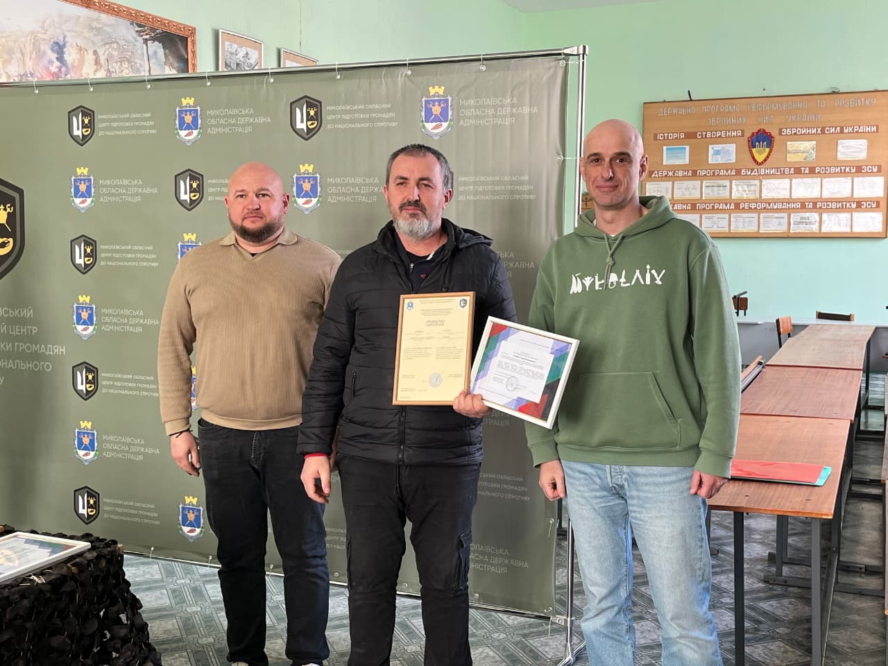 Троє чоловіків у приміщенні на фоні офіційного банера з гербами та інформаційного стенда. Чоловік у центрі — з темним волоссям та сивиною на бороді, одягнений у чорну куртку, тримає в руках дві почесні грамоти в рамках. Поряд з ним стоять двоє чоловіків у повсякденному одязі (світлий джемпер та зелене худі). Атмосфера події — урочисте нагородження.