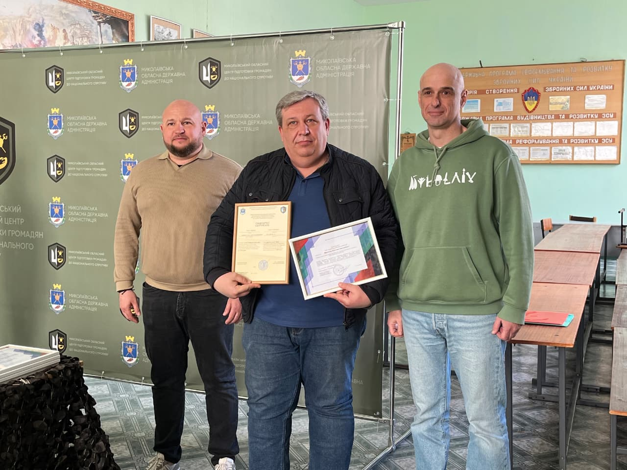 Троє чоловіків у приміщенні на фоні офіційного банера з гербами та інформаційного стенда. Чоловік у центрі — міцної статури, з сивим волоссям, одягнений у синю футболку та чорну куртку, тримає в руках дві почесні грамоти в рамках. Поряд з ним стоять двоє чоловіків у повсякденному одязі (світлий джемпер та зелене худі). Атмосфера події — урочисте нагородження.