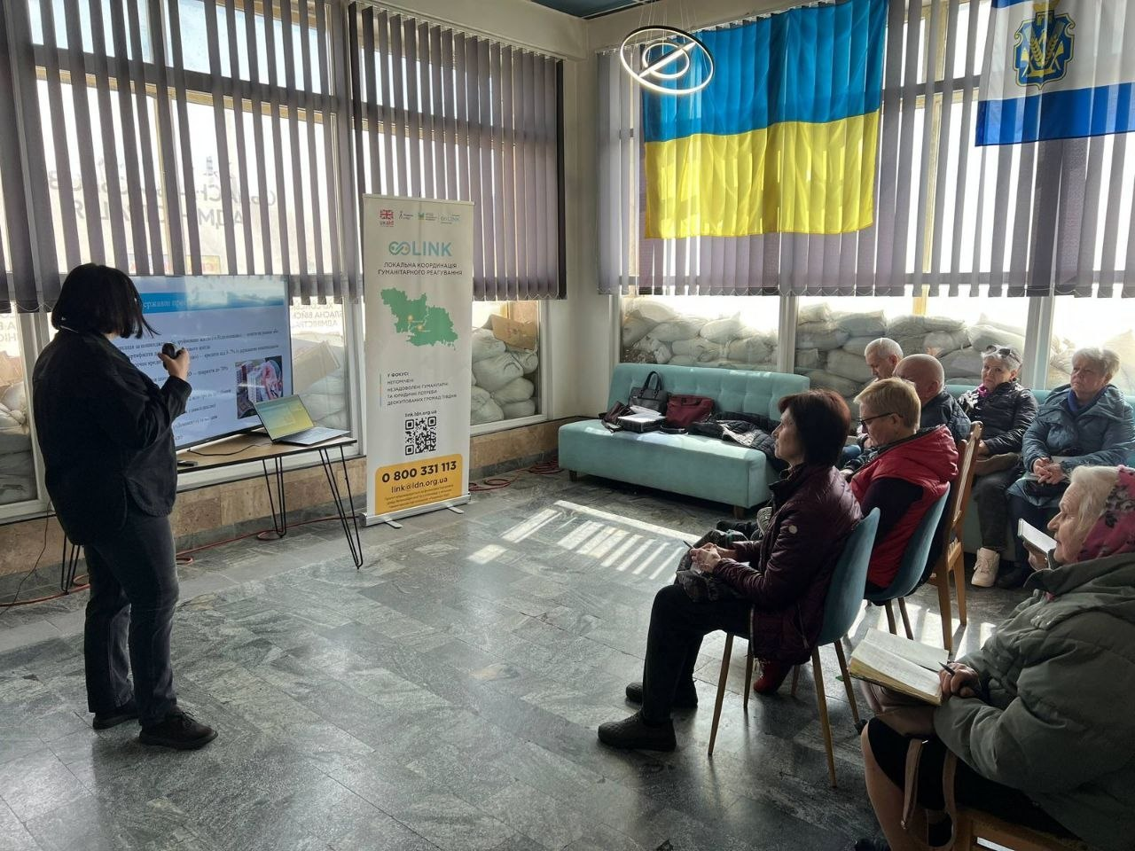 Жінка проводить презентацію для групи людей у світлому залі з українською символікою та вікнами, захищеними мішками з піском. Поруч стоїть інформаційний стенд організацій «Lumos» та «GOLINK».