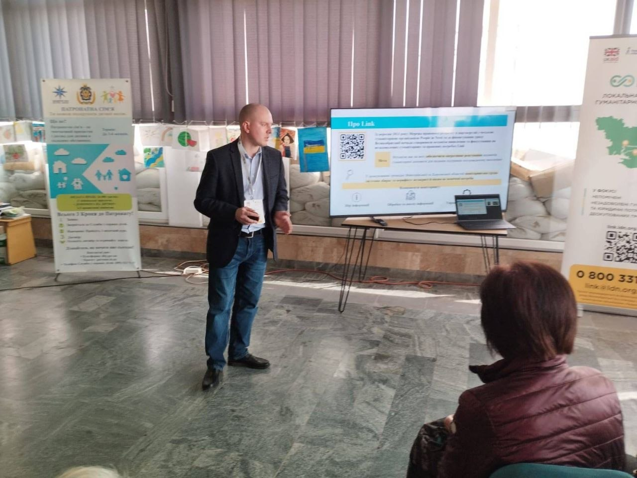 Чоловік проводить презентацію в залі, де вікна захищені мішками з піском. На екрані та банерах поруч розміщена інформація про гуманітарні ініціативи, зокрема проєкт «GoLink» та організацію «Lumos».