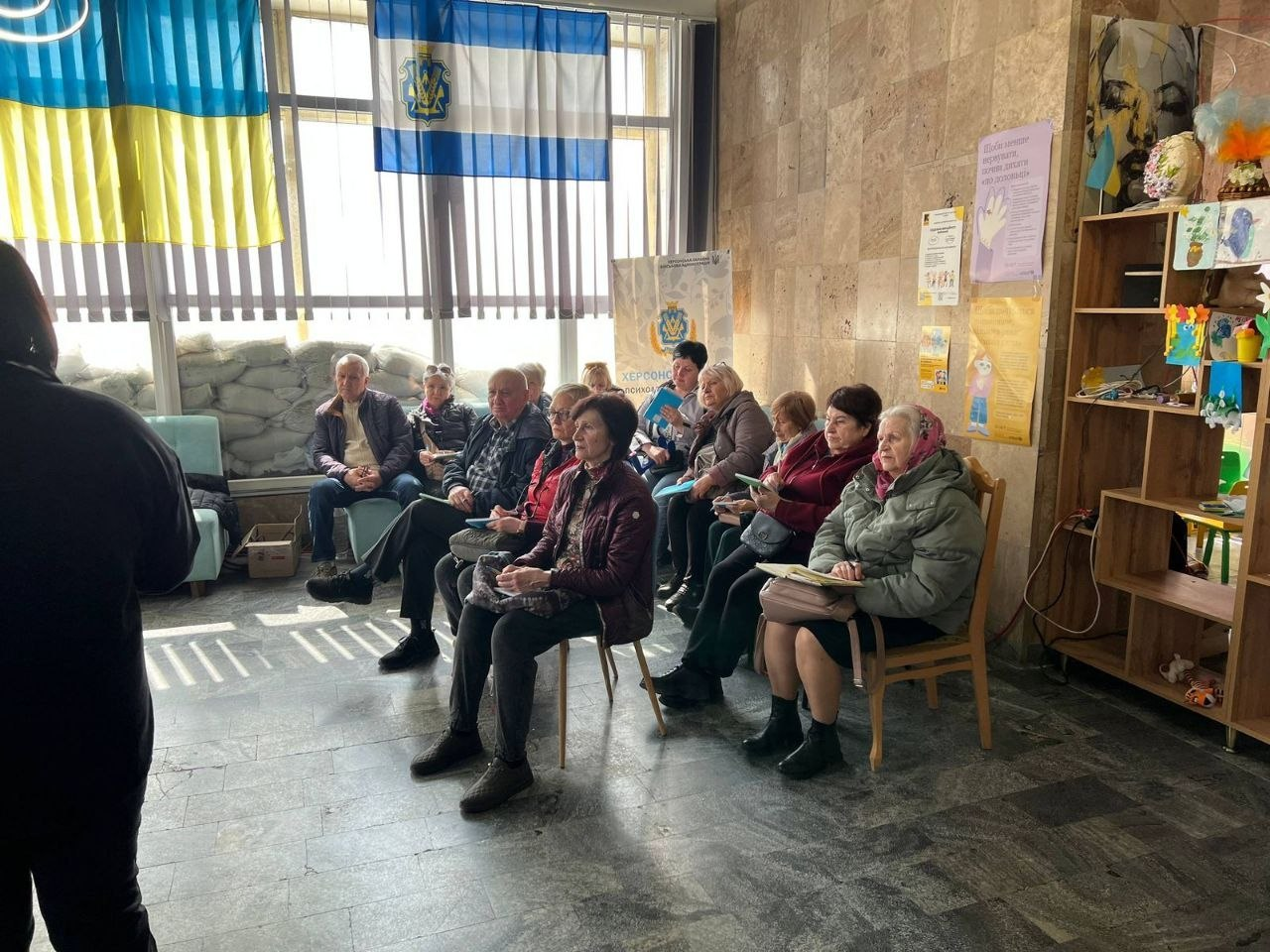 Група людей похилого віку сидить у залі під час заходу. На великому вікні висить прапор України, а підвіконня закладене мішками з піском для захисту.