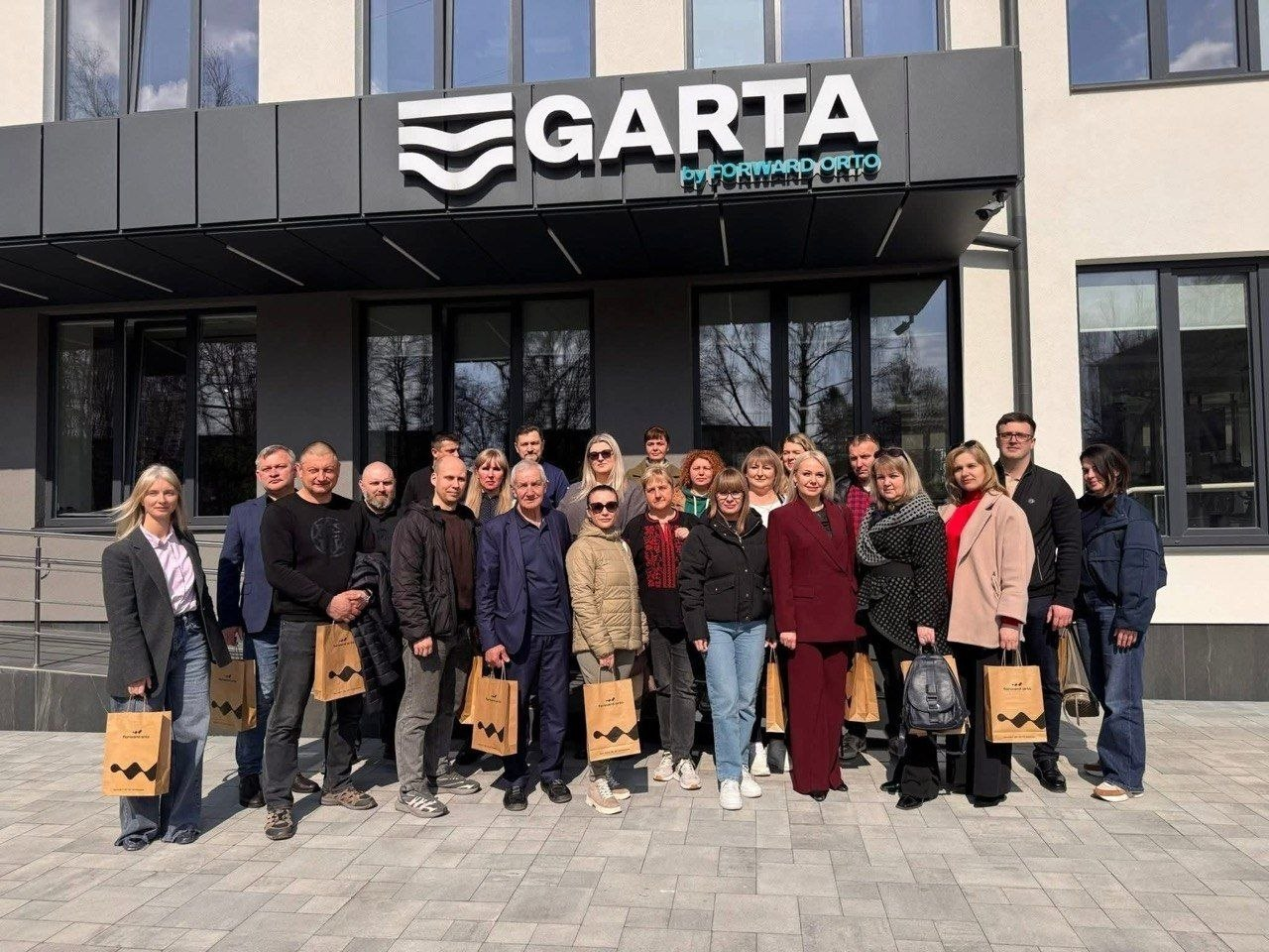 Група людей позує для спільного фото перед сучасним офісним центром під вивіскою «GARTA». Більшість присутніх тримають у руках паперові брендовані пакети.
