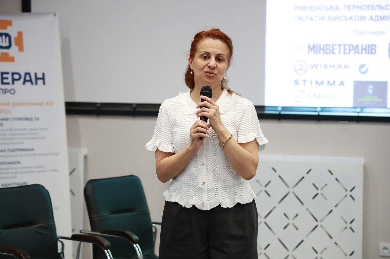 Жінка виступає з мікрофоном на офіційному заході. На фоні видно інформаційний банер та екран із логотипами партнерів, серед яких Міністерство у справах ветеранів України.