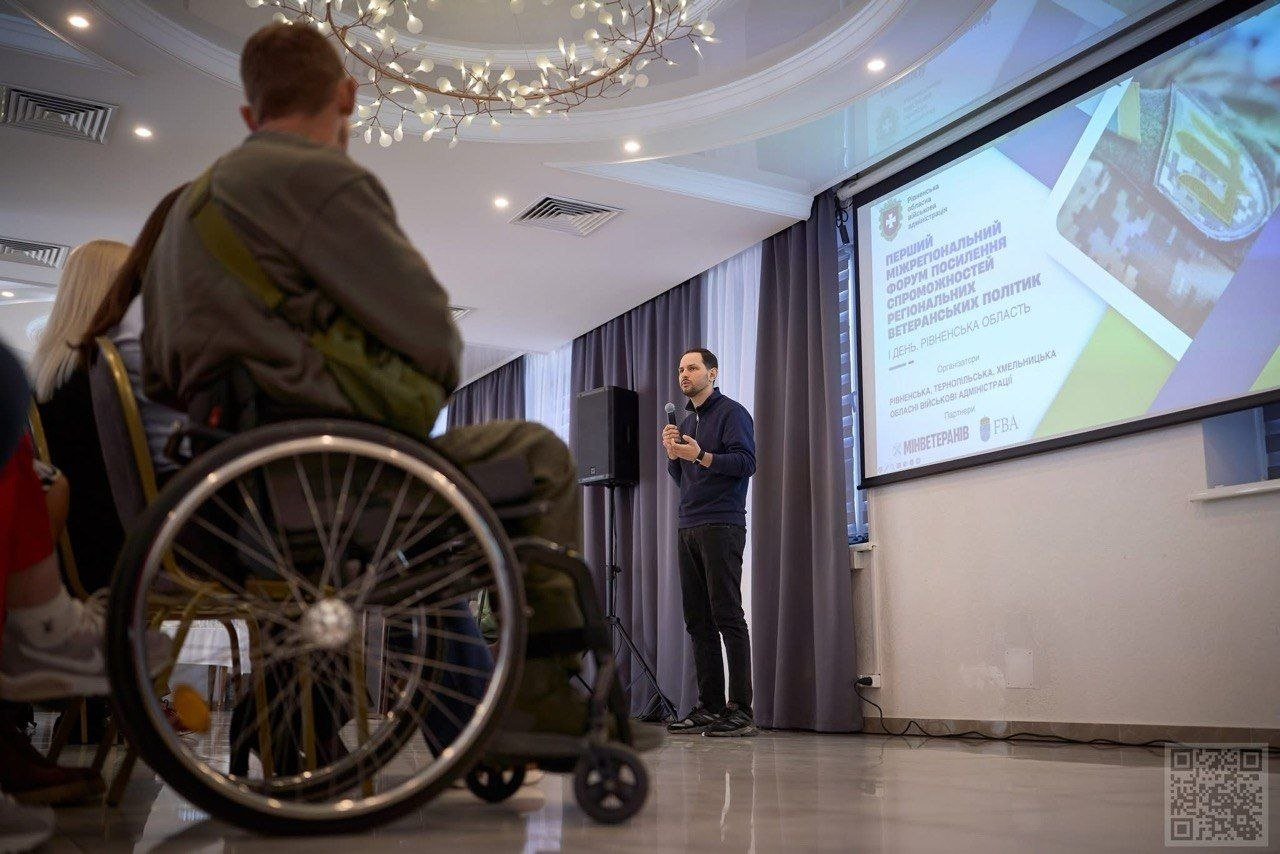 Чоловік виступає перед аудиторією на форумі з посилення регіональних ветеранських політик у Рівненській області. На екрані поруч відображено слайд із деталями заходу та логотипами організаторів, а серед слухачів на передньому плані видно людину на кріслі колісному.